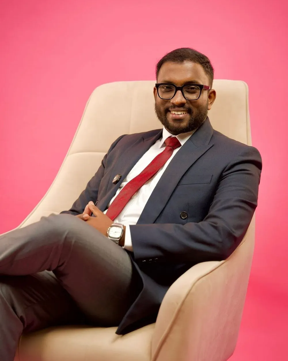 Mayantha seated in a cream armchair against a pink studio backdrop, in a navy suit and red tie