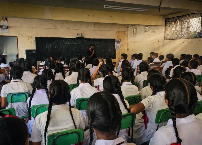 Mayantha Warnakulasooriya conducting a career guidance workshop on stage or in a school auditorium, holding a microphone in professional attire while engaging with an audience of high school students and teachers.